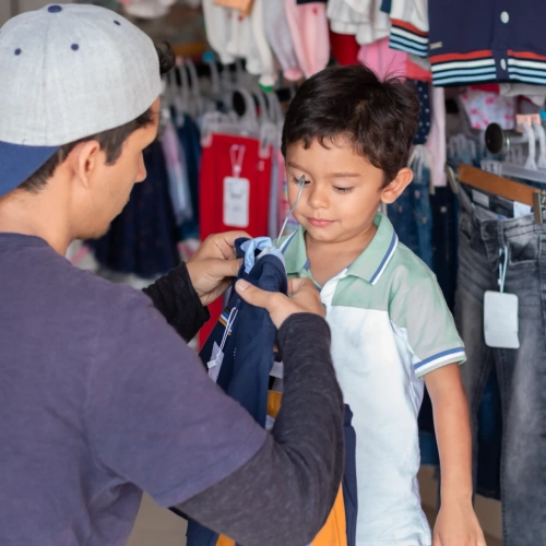 Un homme aide un jeune garçon à essayer des vêtements seconde main enfant dans un magasin, en tendant une chemise à l'enfant alors qu'il est entouré d'étagères de vêtements. Le garçon reste immobile, regardant la chemise.