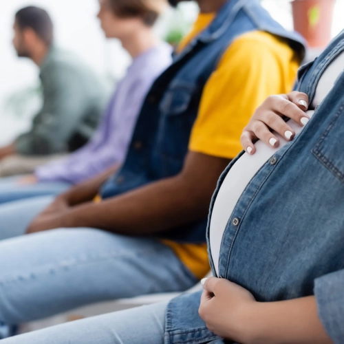 Une personne enceinte vêtue d'une chemise en jean est assise dans une rangée avec d'autres personnes dans un atelier grossesse, posant sa main sur son baby bump. L'accent est mis sur le torse de la personne enceinte, les autres personnes étant floues à l'arrière-plan.
