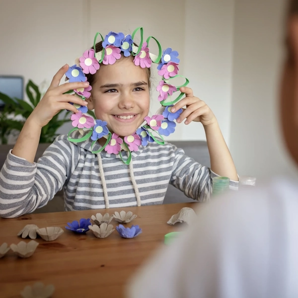 Un enfant souriant portant un objet diy en forme de fleur autour de son visage est assis à une table avec du matériel de bricolage, tandis qu'un autre enfant lui fait face au premier plan.
